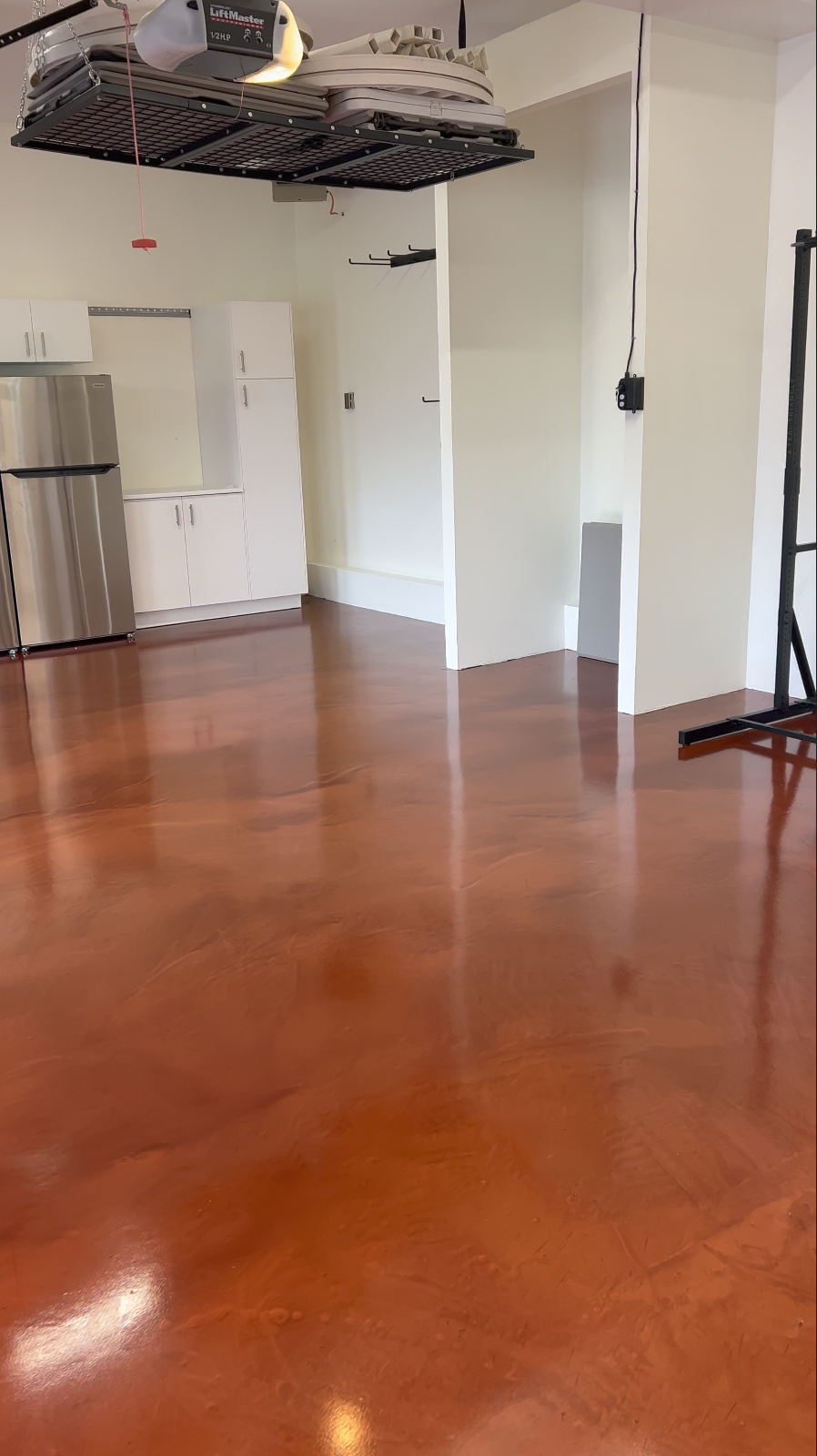 Empty room with polished reddish-brown hardwood floors, white walls, white cabinets, a stainless steel refrigerator, and a garage door opener system hanging from the ceiling.