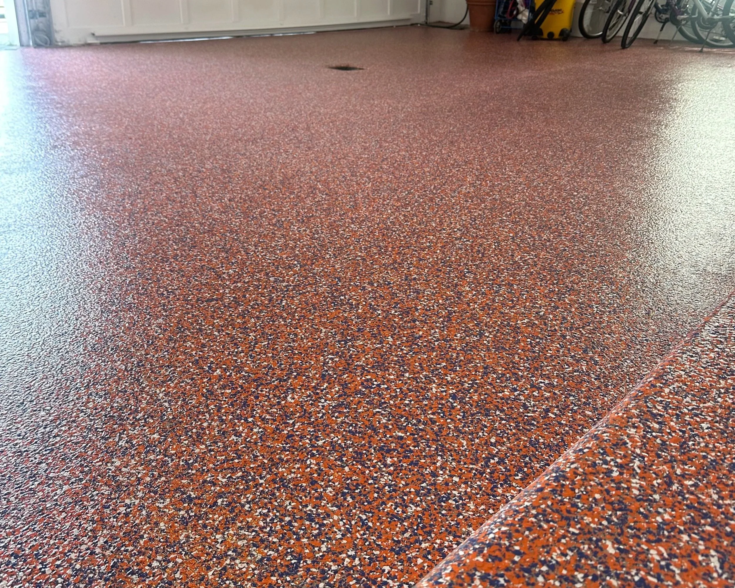 Close-up of a speckled red, black, and white epoxy flooring in a garage, with garage doors visible in the background and a few items stored along the walls.