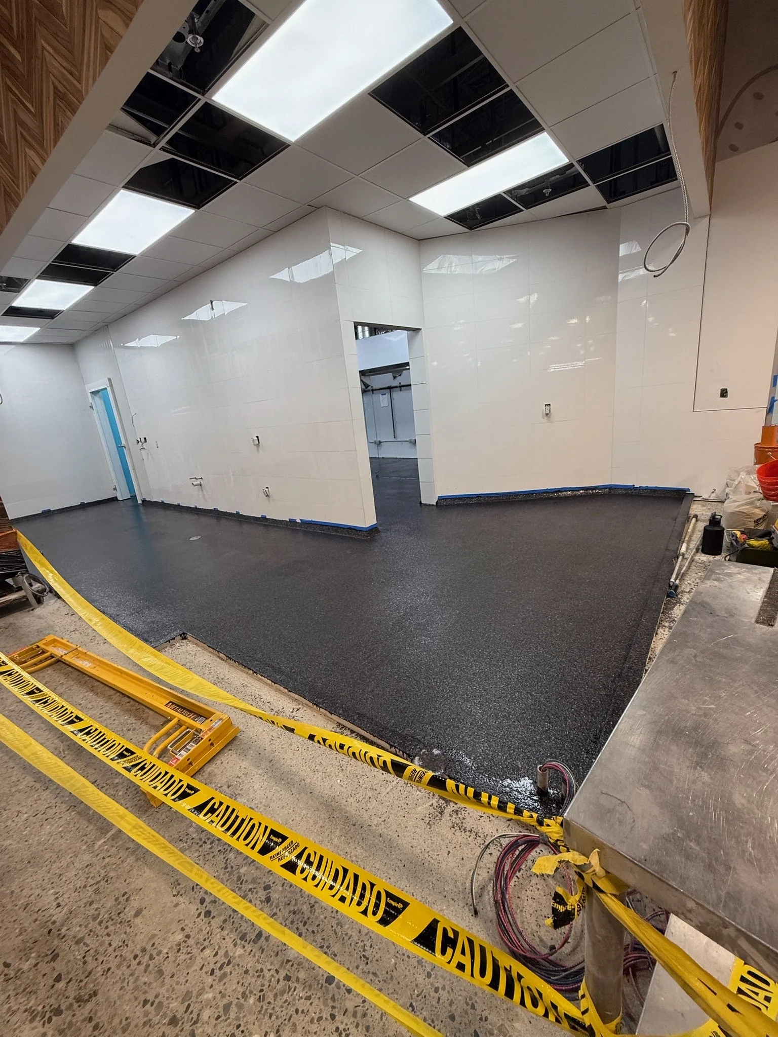 Construction site with partially finished ceiling and walls. The ceiling has white and black panels, some of which are missing. The floors are uncovered, and caution tape surrounds the work area.
