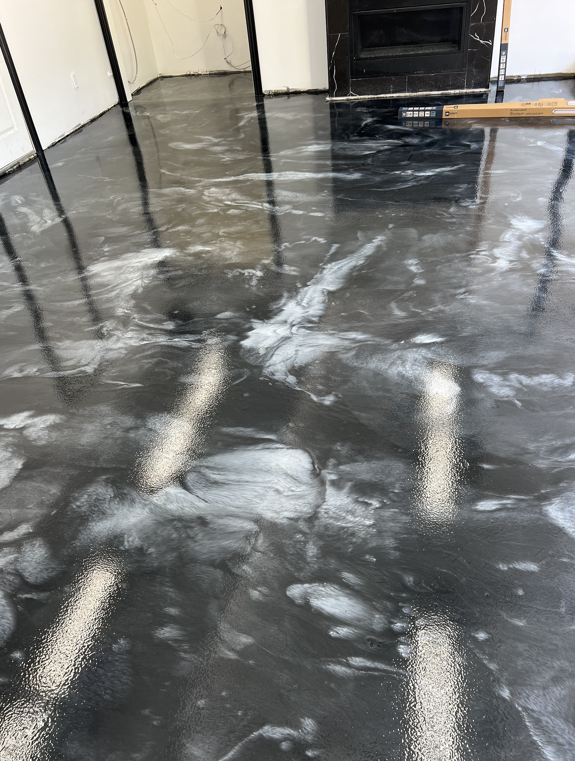 A room with a concrete floor that is wet and shiny, with reflections of walls and ceiling lights. The room has white walls, some black metal supports, and a black fireplace or fireplace surround area. There are some construction materials and wires v