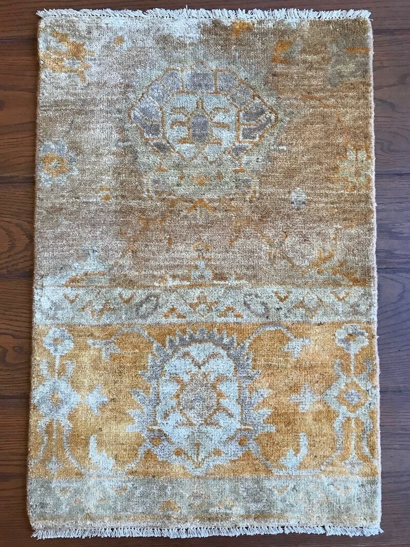 A rectangular area rug with a traditional ornate pattern in shades of beige, gold, and light gray on a wooden floor.