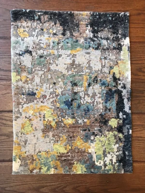 A rectangular area rug with an abstract, multicolored pattern featuring shades of black, gray, yellow, green, blue, and beige, placed on a wooden floor.