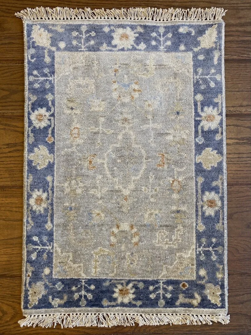 A small rectangular area rug with a floral and geometric pattern, featuring a blue border with white and beige flowers, on a wooden floor.