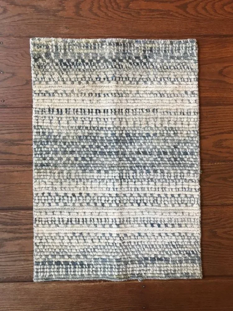 A rectangular, woven area rug with a neutral, striped pattern in shades of cream, gray, and black, placed on a wooden floor.