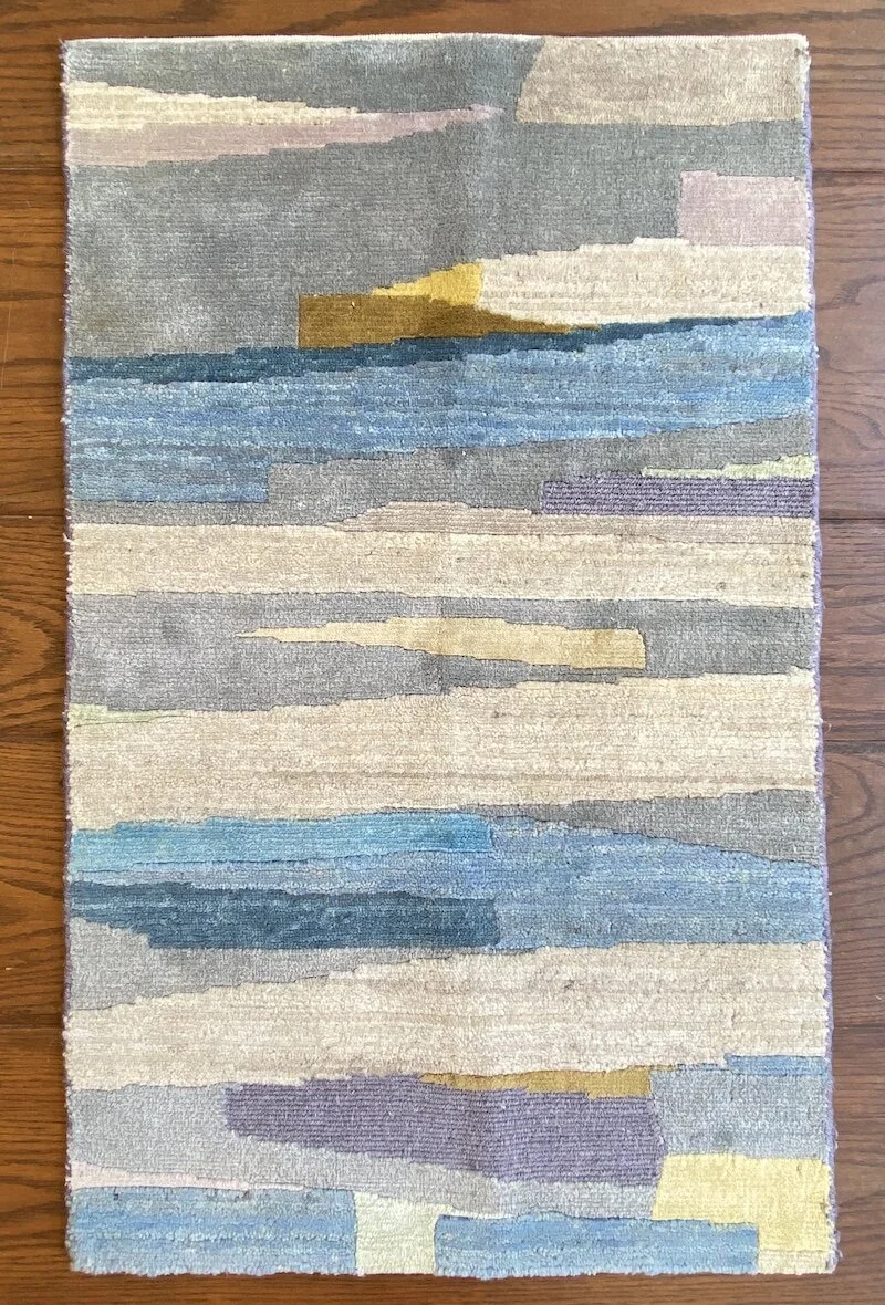A rectangular abstract rug with horizontal bands in shades of blue, gray, beige, yellow, and purple, on a wooden floor.