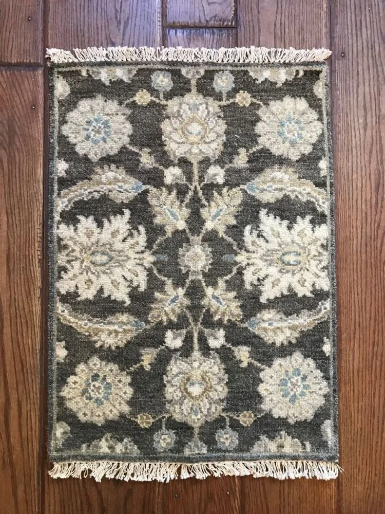 Small decorative rug with a floral pattern in beige, cream, and light blue colors on a dark background, with fringed edges.