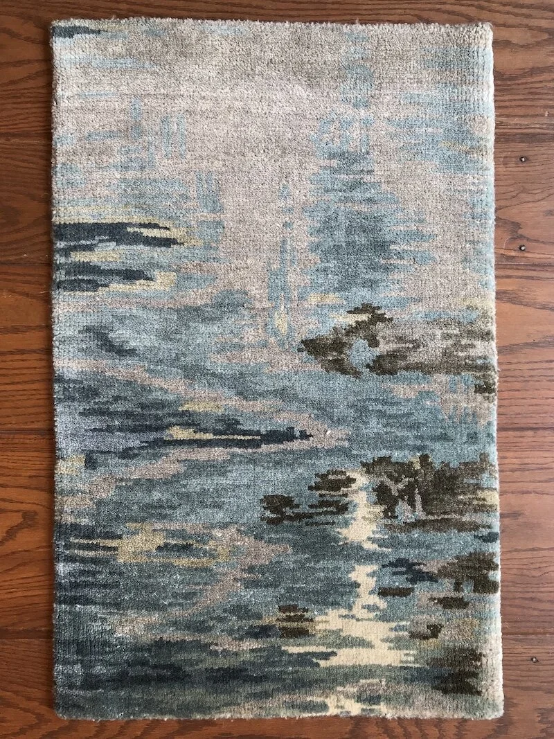 A rectangular area rug with a landscape scene of trees and water reflected in a calm lake, in shades of blue, beige, and black, placed on a wooden floor.