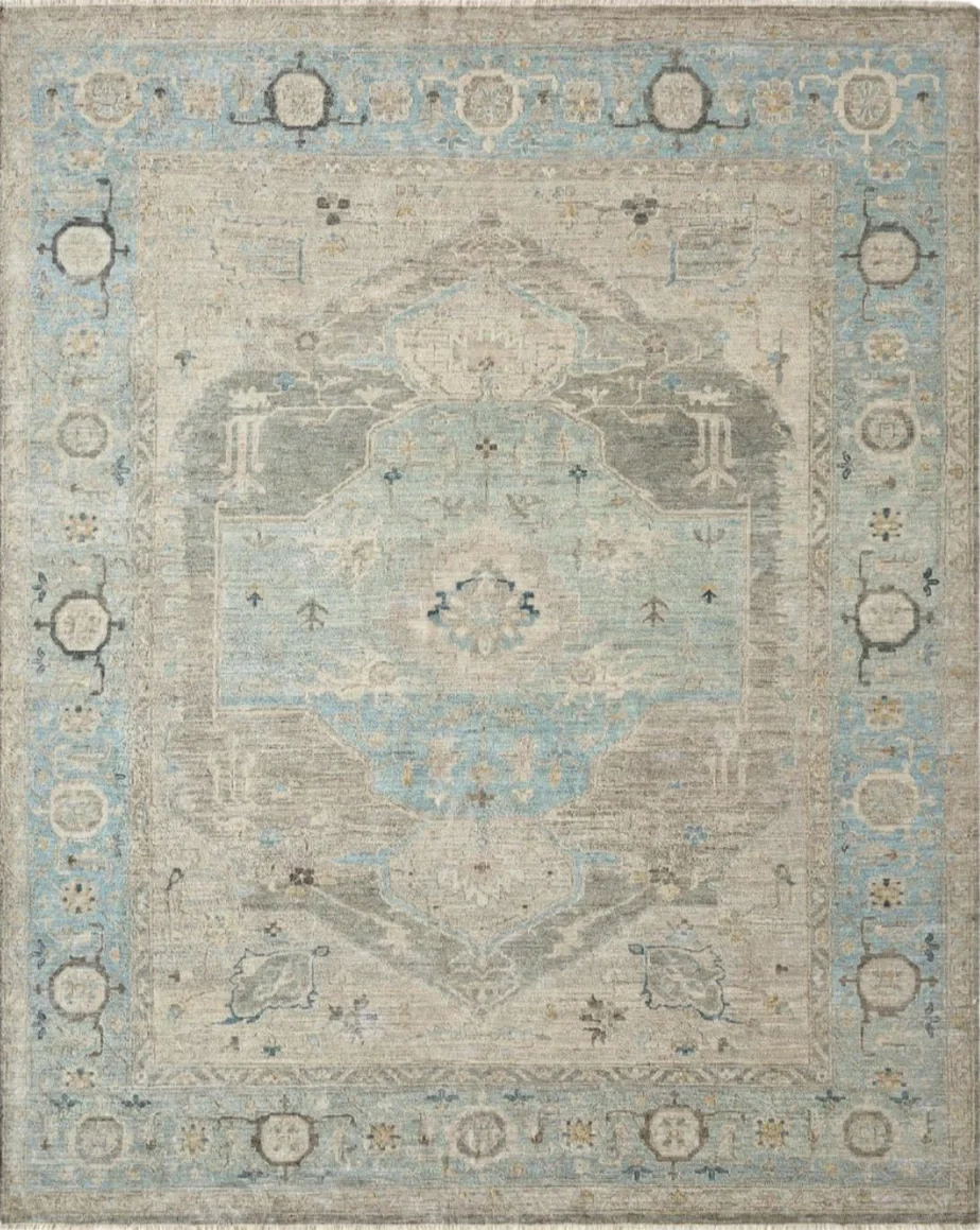 A vintage, faded area rug with a central medallion design in cream, light gray, and pale blue hues, featuring a geometric border pattern.