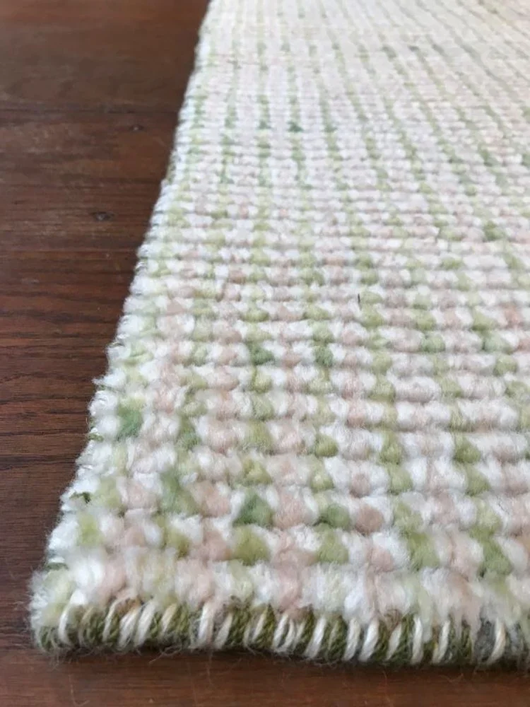 Close-up of a woven area rug with white, green, and peach-colored yarn, placed on a hardwood floor.