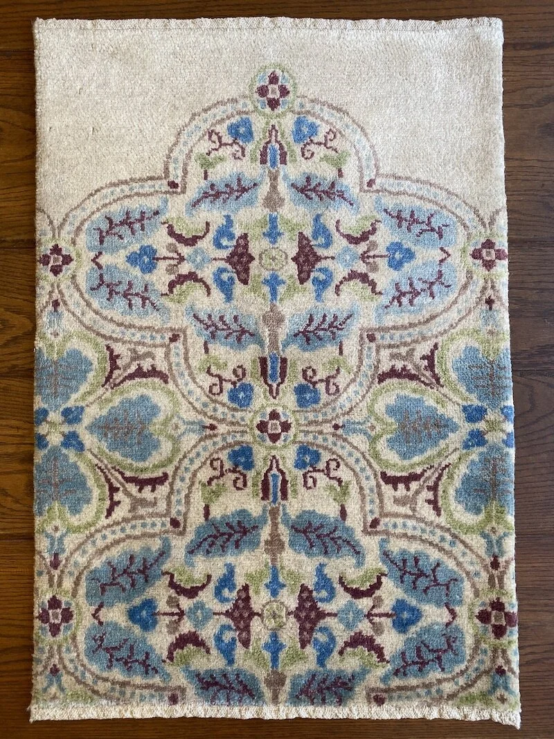 A decorative area rug with a symmetrical floral and leaf pattern in shades of blue, green, burgundy, and beige on a wooden floor.