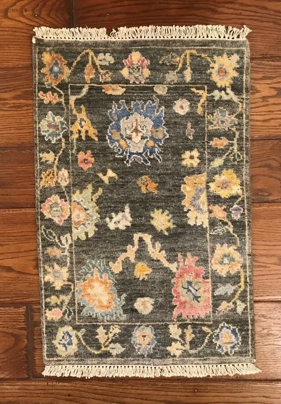 Small decorative rug with a dark background, floral patterns in pastel colors, and fringes on both sides, laid on a wooden floor.