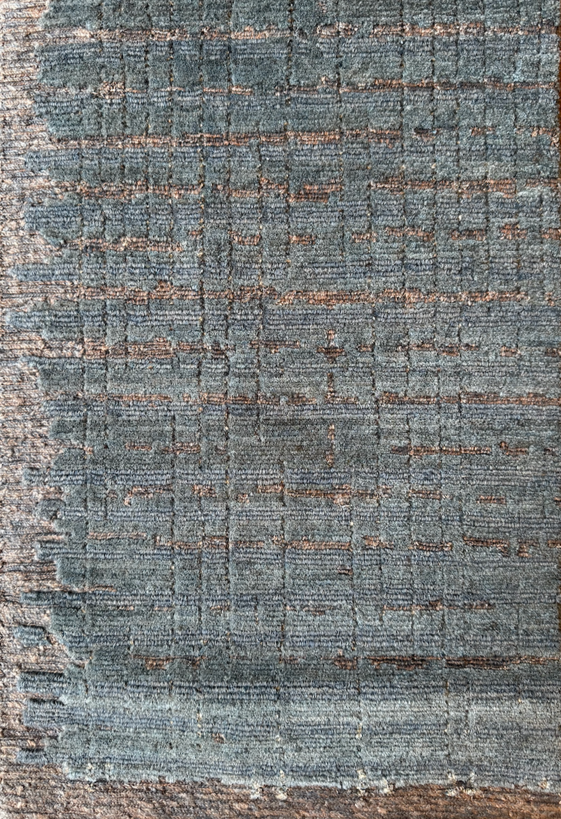 Close-up of a textured, woven fabric or carpet with a pattern of intersecting lines and rectangles in shades of blue, brown, and gray.