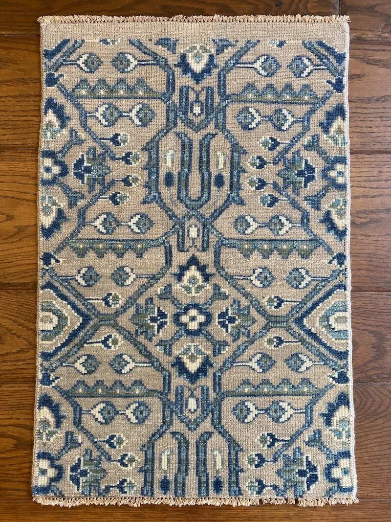 A rectangular woven rug with intricate geometric and floral patterns in shades of blue, beige, and white, placed on a wooden floor.