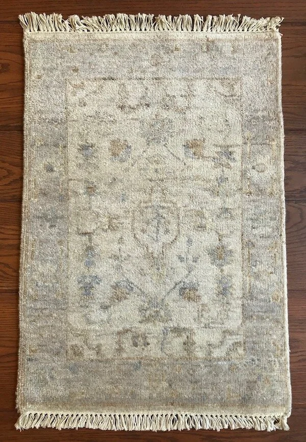 A beige and light gray patterned area rug with fringe along the top and bottom edges, laid on a wooden floor.