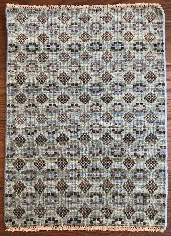 A rectangular area rug with a geometric pattern of diamond and oval shapes in various shades of blue, brown, and beige, with fringed edges on two sides, placed on a wooden floor.