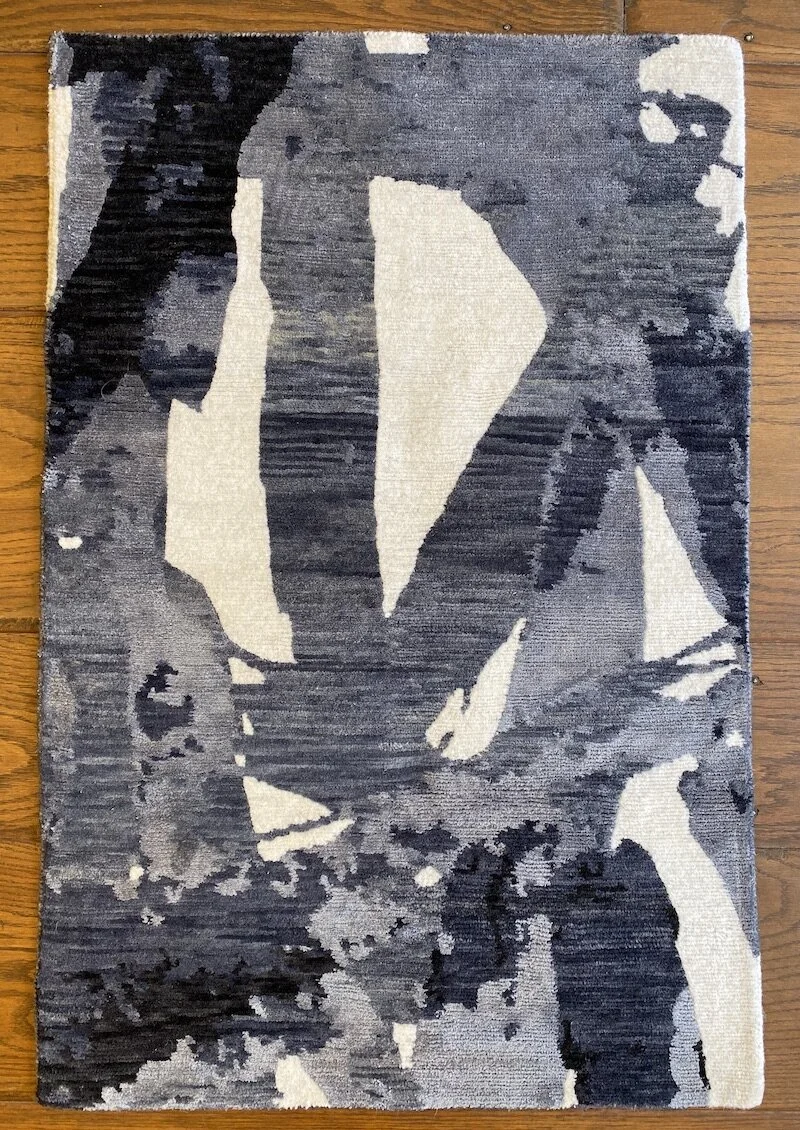 A rectangular patterned rug with abstract black, white, and shades of gray shapes on a wooden floor.