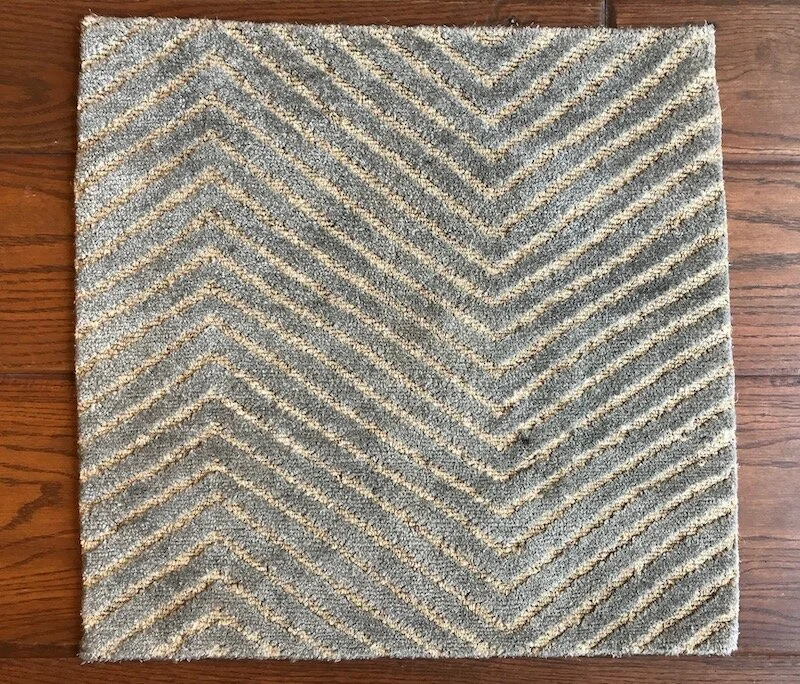 A square gray and beige striped area rug with a chevron pattern on a wooden floor.
