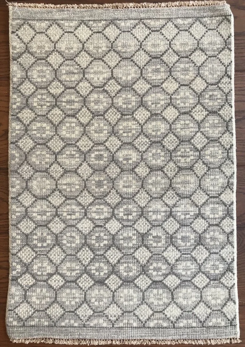 A rectangular area rug with a geometric pattern of interconnected circles and squares in shades of gray and beige, on a wooden floor.