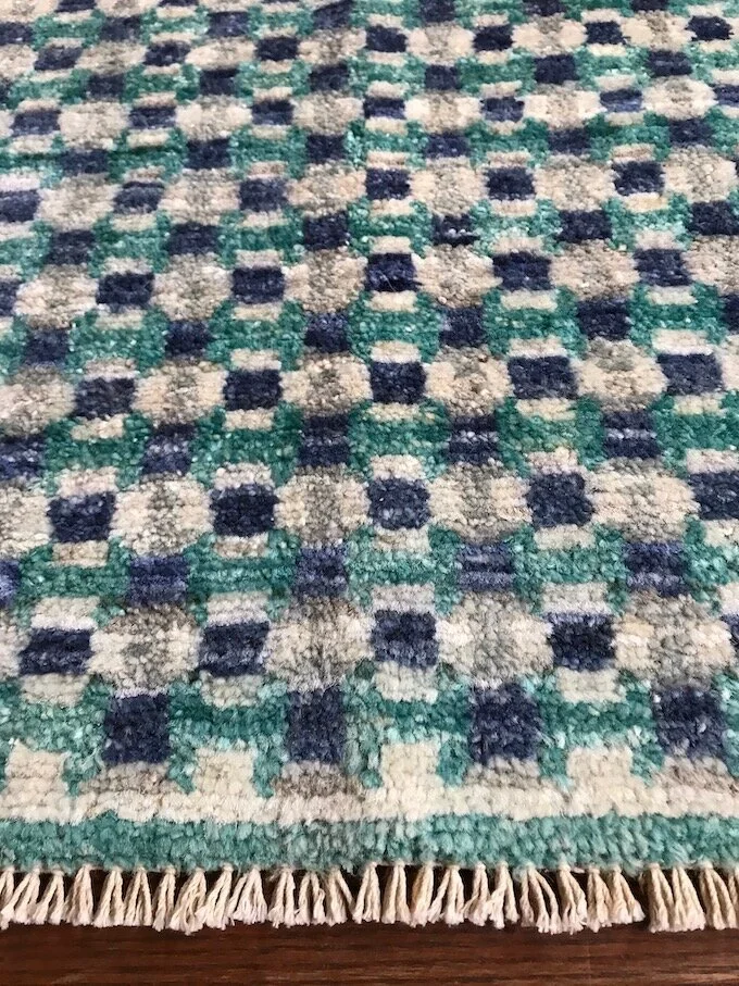 Close-up of a patterned area rug with blue, green, beige, and navy squares and tassels on the edge.
