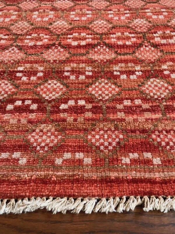 Close-up of a vintage woven rug with a geometric floral pattern in shades of red, pink, white, and green, with fringed edges.