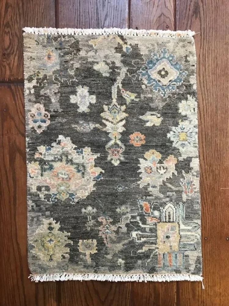A small rectangular area rug with intricate floral and geometric patterns in shades of gray, cream, blue, and orange, with fringe on both ends, placed on a wooden floor.