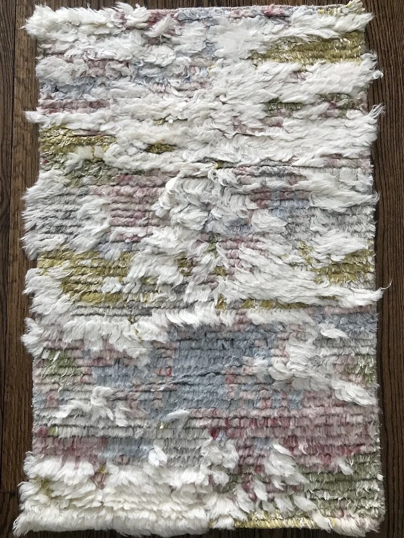 A small, rectangular rug made of layered, shredded fabric strips in white, pink, yellow, and blue on a wooden floor.