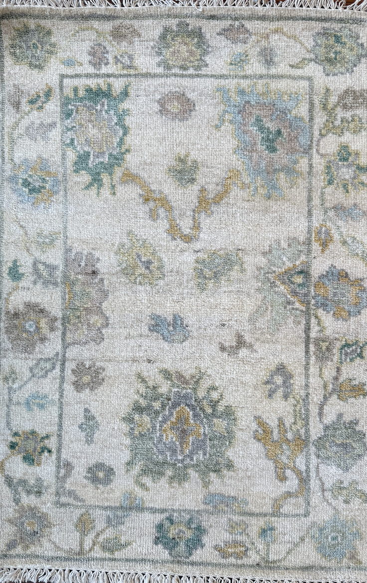 An antique floral patterned area rug with cream, light blue, gray, and gold hues and fringed edges.