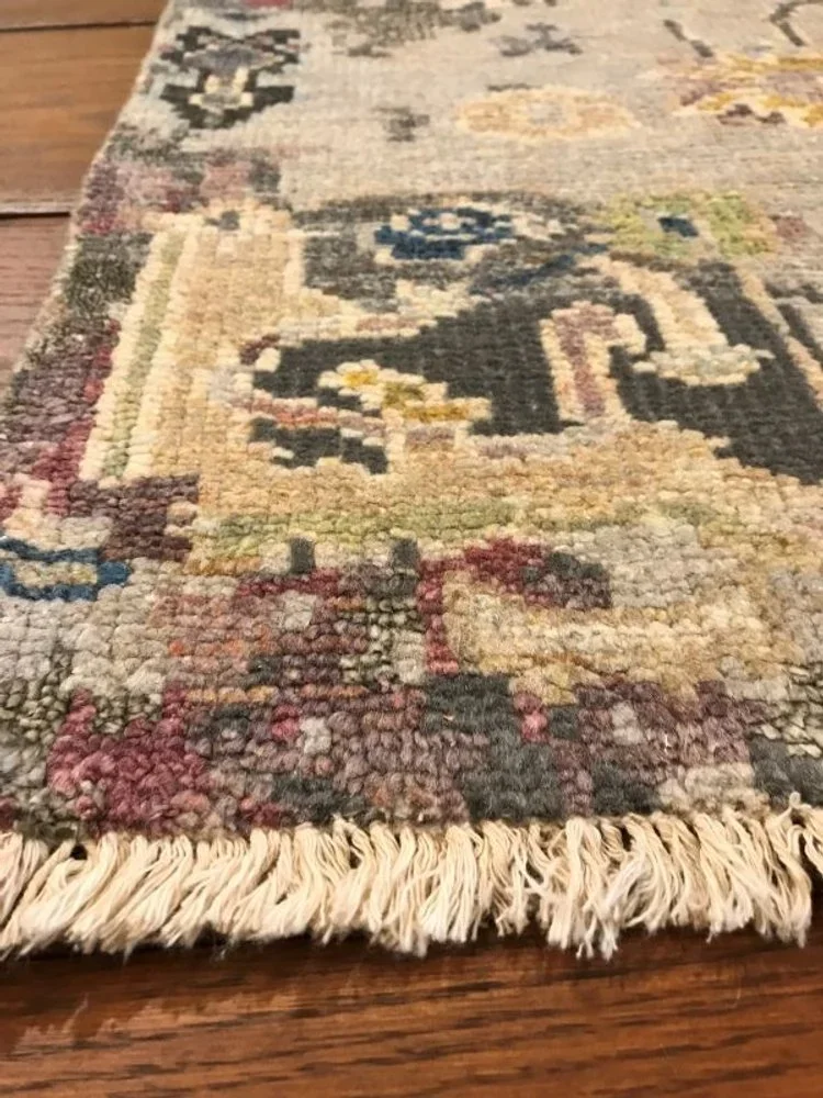 Close-up of a traditional woven area rug with a floral and geometric pattern, fringed edge, on a wooden floor.