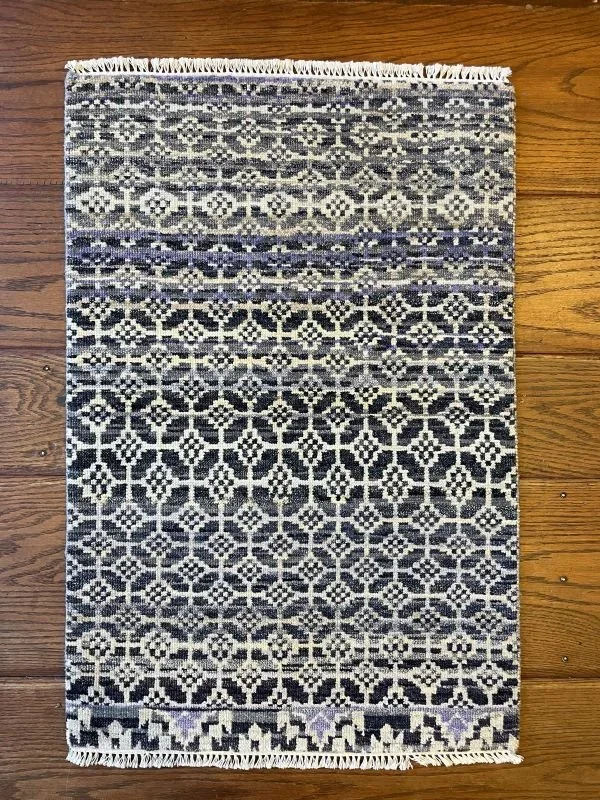 A rectangular woven rug with a geometric black and white pattern, fringes on both shorter sides, placed on a hardwood floor.