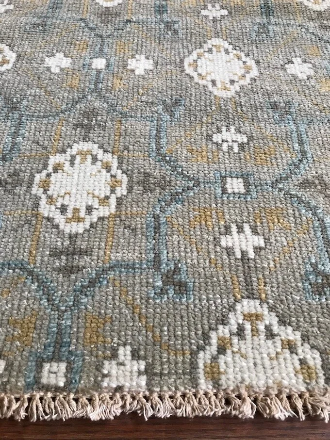 Close-up of a decorative area rug with a floral and geometric pattern in neutral colors, featuring white, gray, beige, and blue accents, with fringe on the edge.