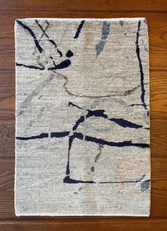 Light beige area rug with abstract dark gray and navy lines and splashes, placed on a wooden floor.