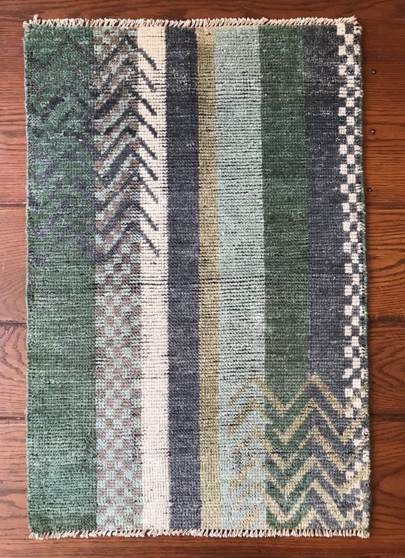 Colorful rectangular woven rug with geometric patterns in green, black, gray, white, and beige, on a wooden floor.