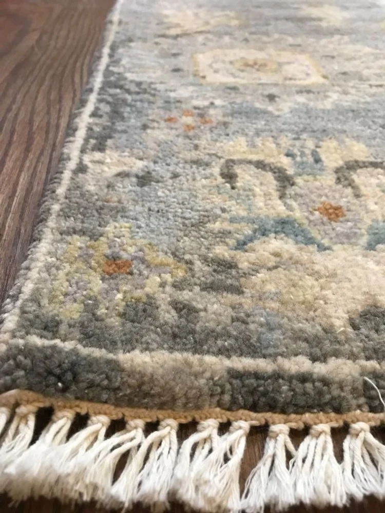 Close-up of a patterned area rug with fringe on the edge, placed on a wooden floor.