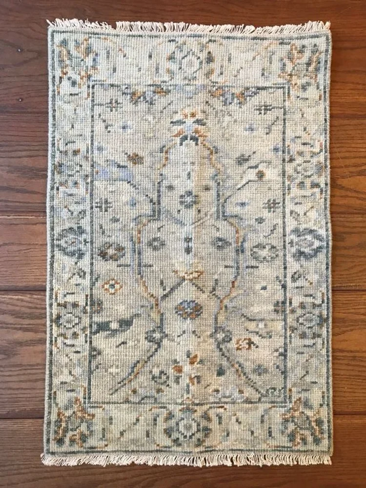 A small, rectangular, light-colored area rug with intricate traditional patterns in muted tones of beige, gray, blue, and brown on a wooden floor.