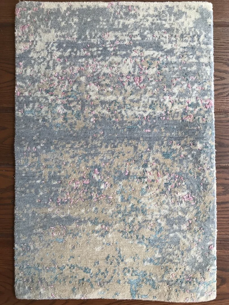 A rectangular area rug with an abstract pattern in shades of blue, beige, and pink, placed on a wooden floor.