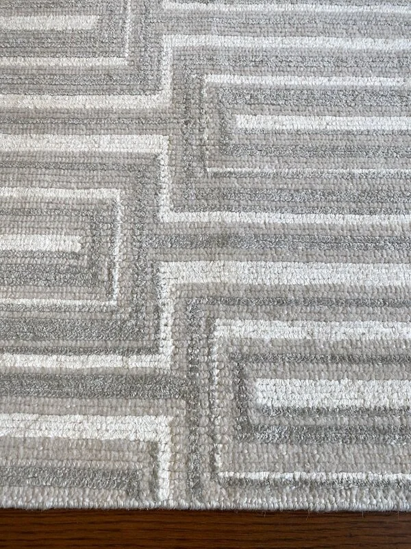 Close-up of a textured, patterned area rug with geometric designs in shades of beige, gray, and white, placed on a hardwood floor.