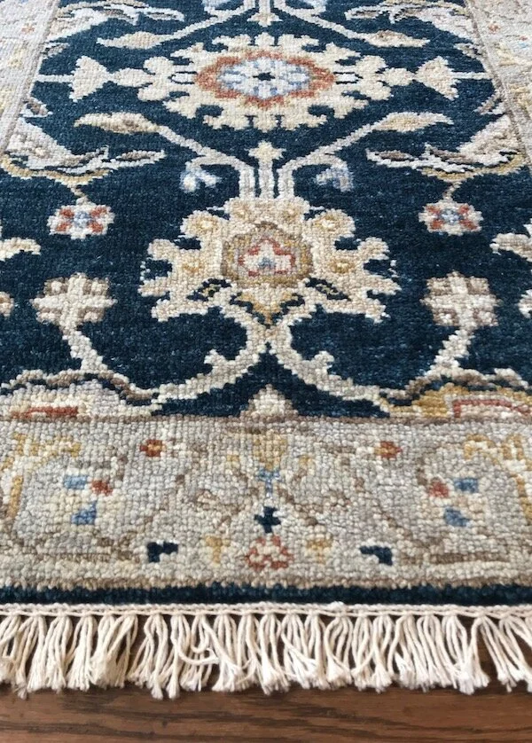 Close-up of a traditional decorative area rug with a floral pattern in beige, navy blue, and light brown, fringed at the bottom edge, on a wooden floor.