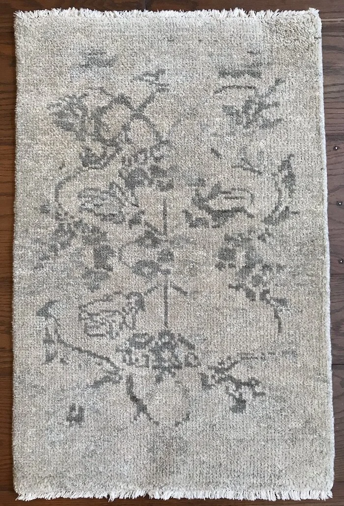 A rectangular beige area rug with a faded floral and vine pattern in gray, placed on a wooden floor.