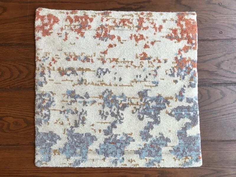 Square patterned area rug with abstract design in shades of blue, orange, and beige on a dark hardwood floor.