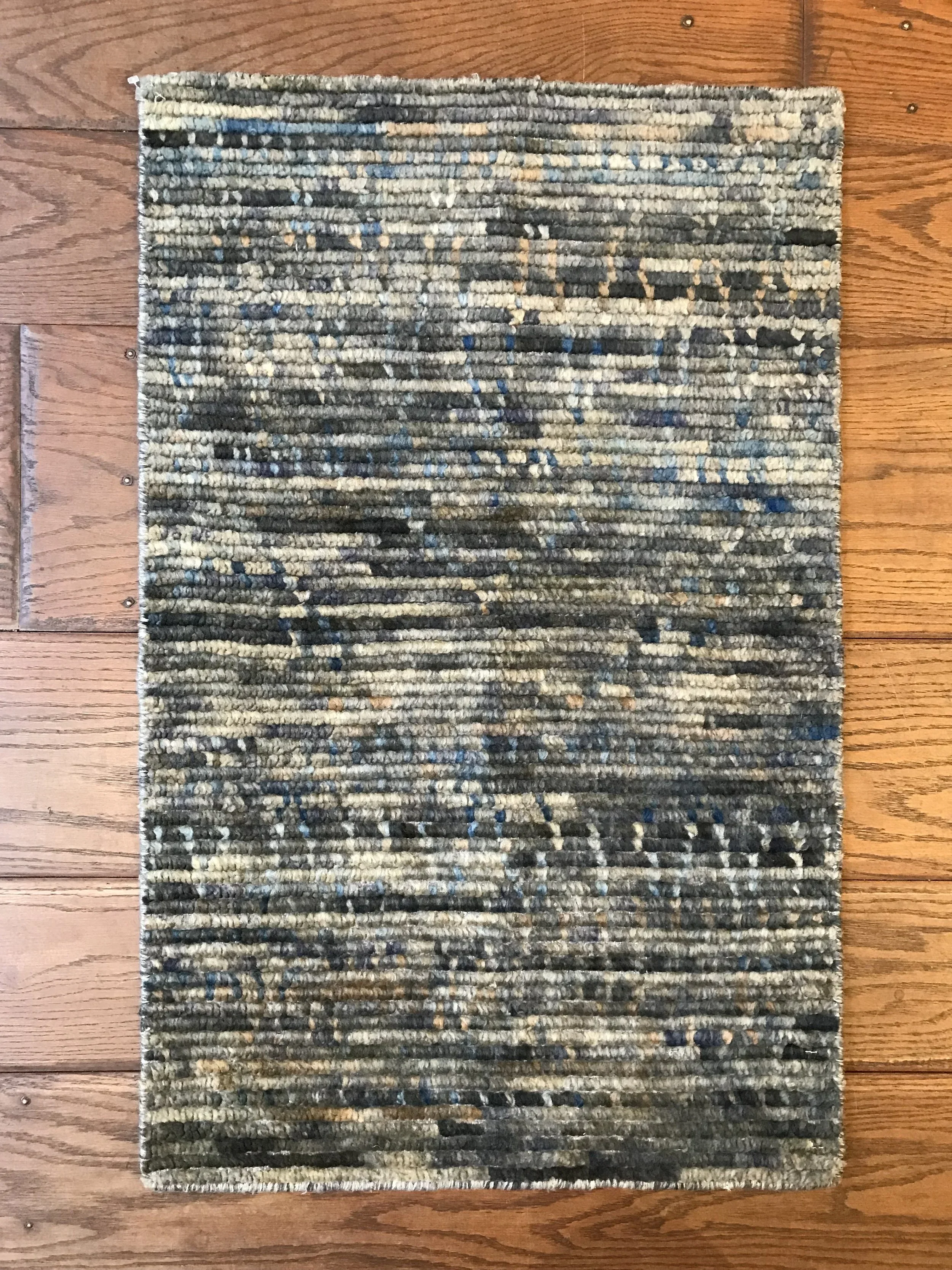 A rectangular, textured area rug with a mix of gray, black, beige, and blue tones, laid out on a wooden floor.