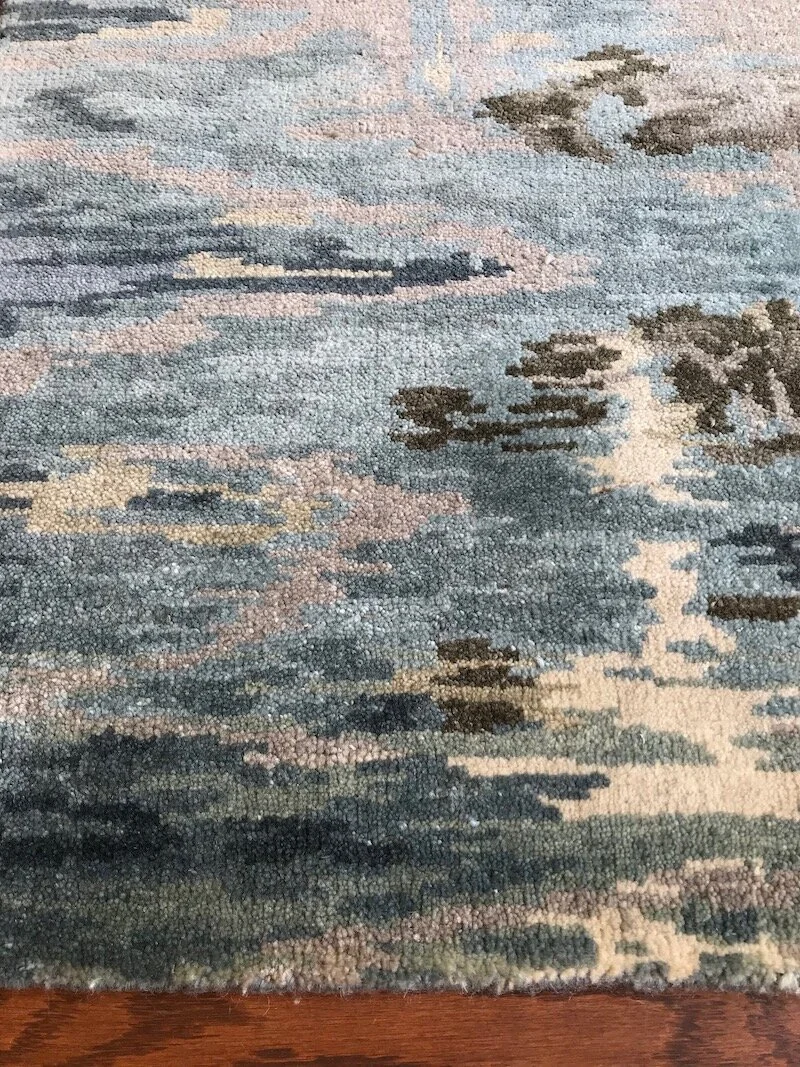 Close-up of an abstract patterned rug with shades of blue, gray, black, cream, and hints of pink, next to a wooden floor.