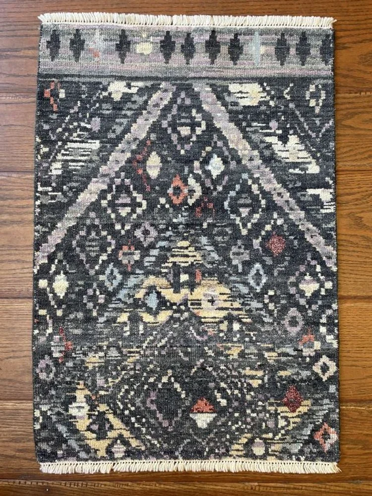 A small dark patterned area rug with geometric designs, fringed borders, placed on a hardwood floor.