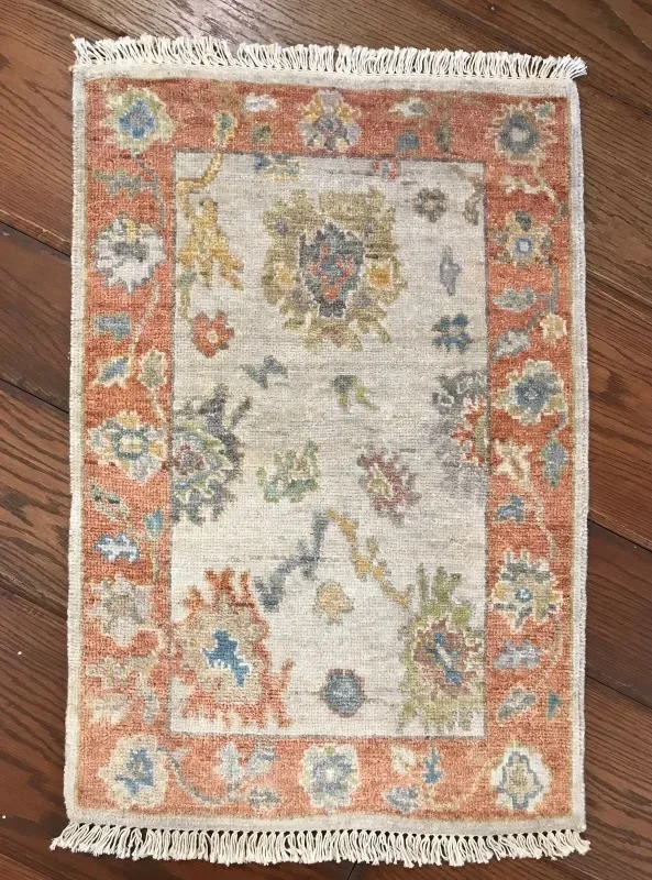 Small multicolored area rug with a floral and geometric pattern, featuring a peach-colored border and fringed edges, placed on a wooden floor.