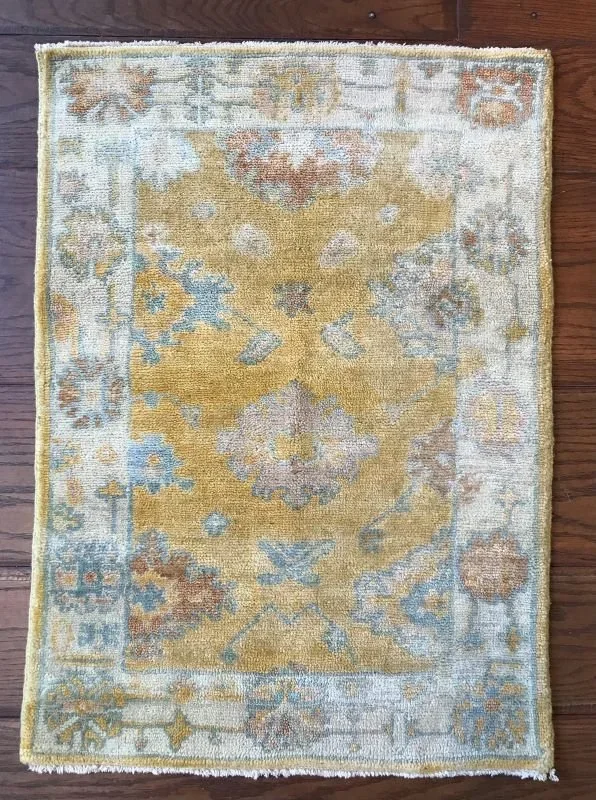 A small vintage rug with a floral and geometric pattern in shades of beige, light blue, and soft pink, on a wooden floor.