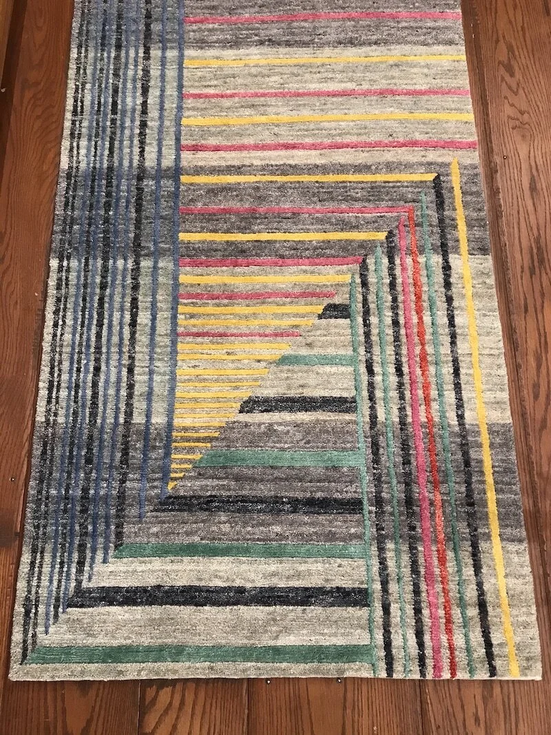 Colorful rectangular area rug with geometric stripe pattern in colors including yellow, red, blue, green, black, and beige, on a wooden floor.