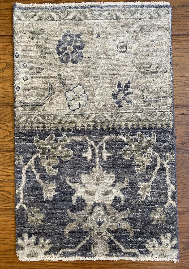 A small, vintage-style area rug with a floral and geometric pattern in shades of beige, gray, and cream, placed on a wooden floor.