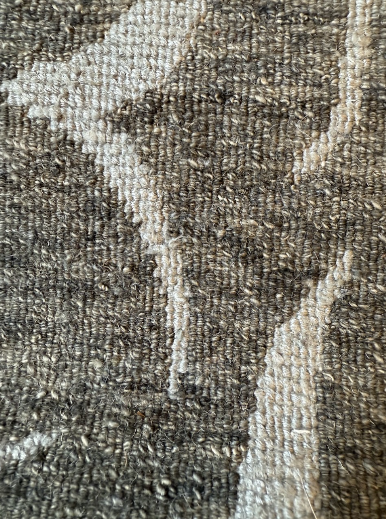 Close-up of a textured, woven fabric with a pattern of darker and lighter yarns, creating a floral or organic design.