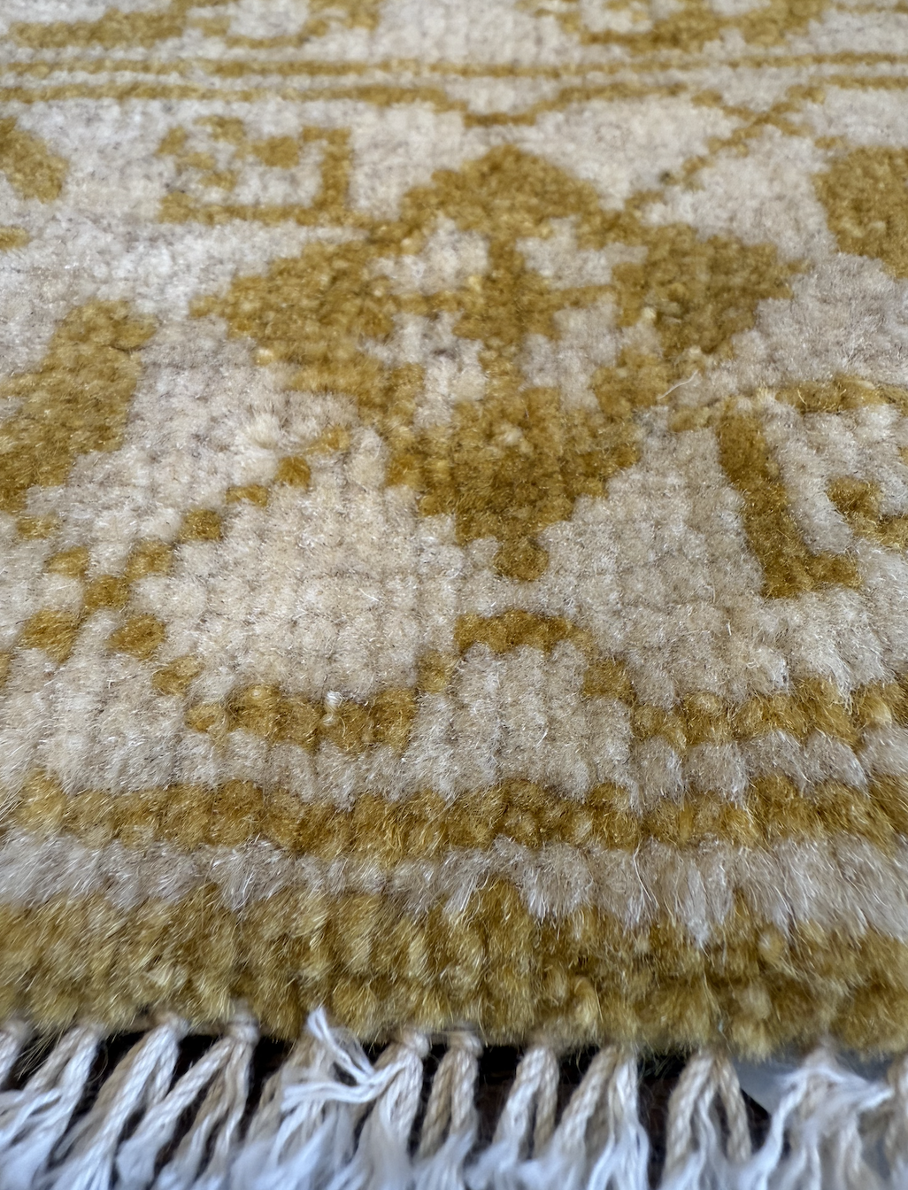 Close-up of a patterned, beige and gold woven carpet with fringed tassels at the bottom.