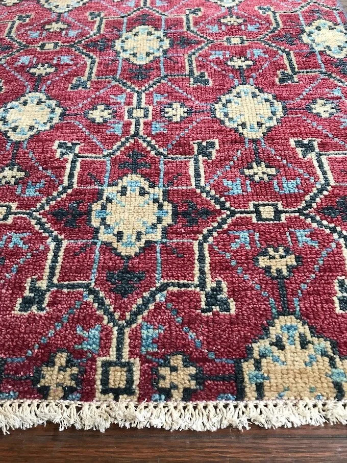 Close-up of a traditional patterned area rug with red, cream, black, and light blue geometric designs and fringed edges on a wooden floor.