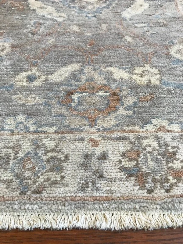 Close-up of a vintage area rug with intricate floral and geometric patterns in muted colors including beige, gray, and light blue, with fringed edges.